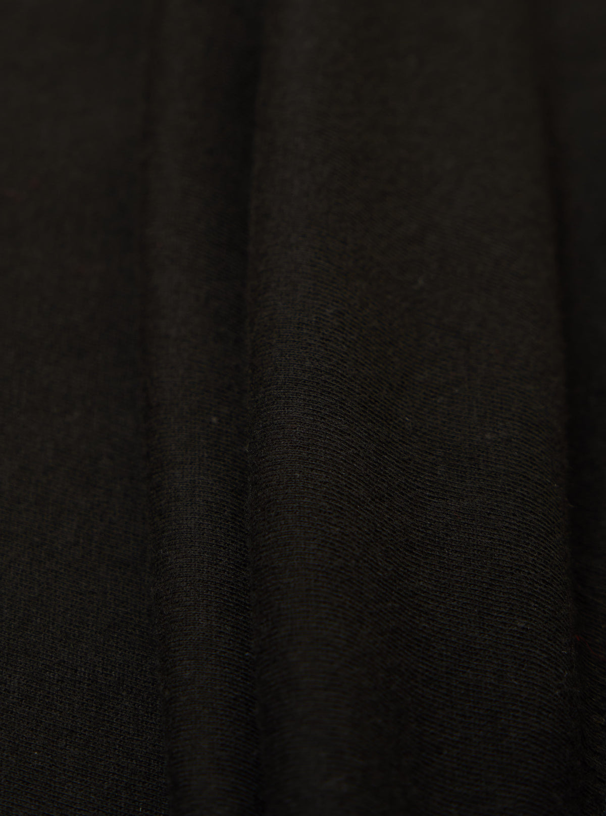 Close-up of the Black Single Jersey Funnel Neck Tee fabric. The soft, black jersey material is draped to show texture. Simple studio background.