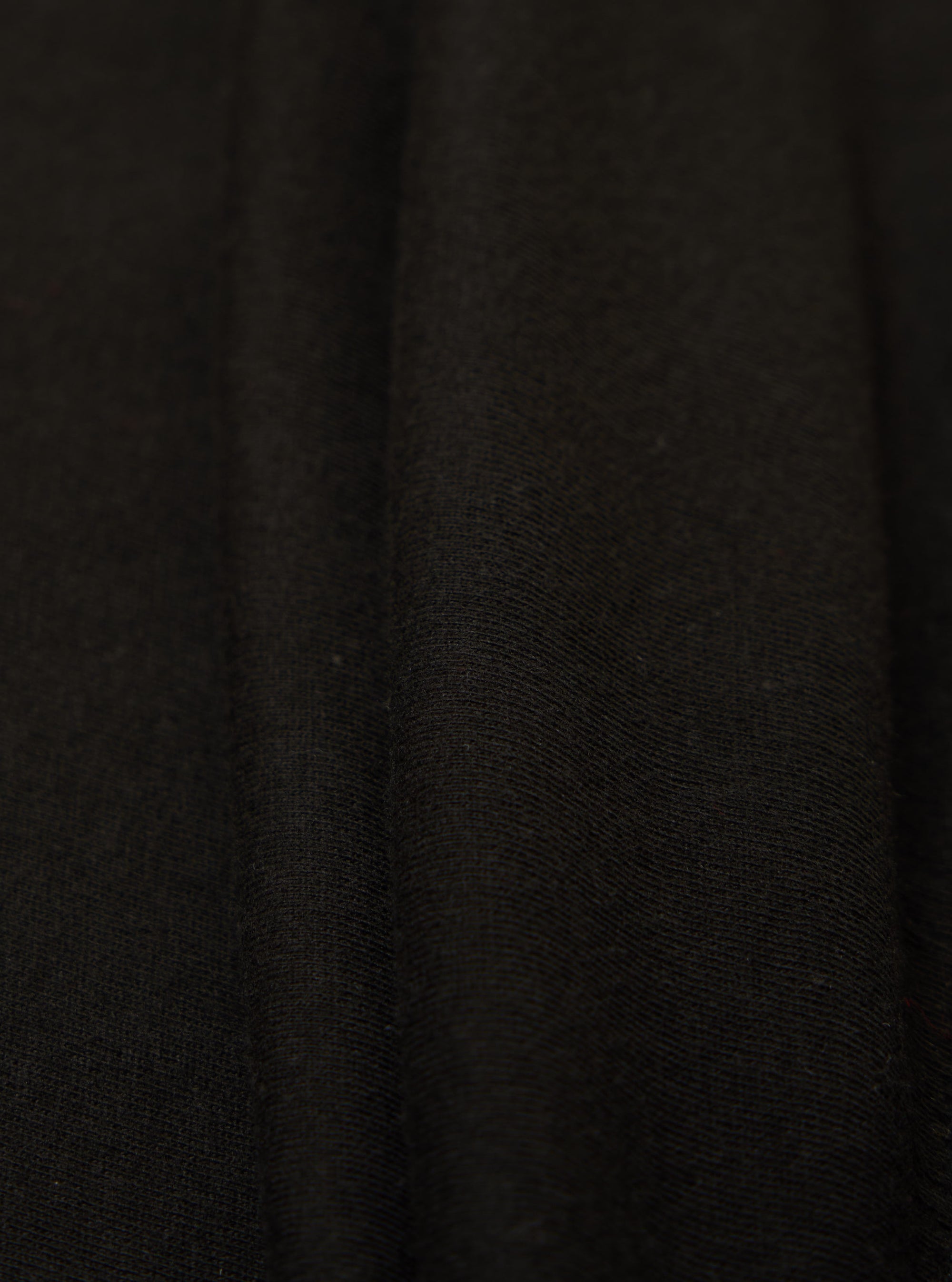 Close-up of the Black Single Jersey Funnel Neck Tee fabric. The soft, black jersey material is draped to show texture. Simple studio background.