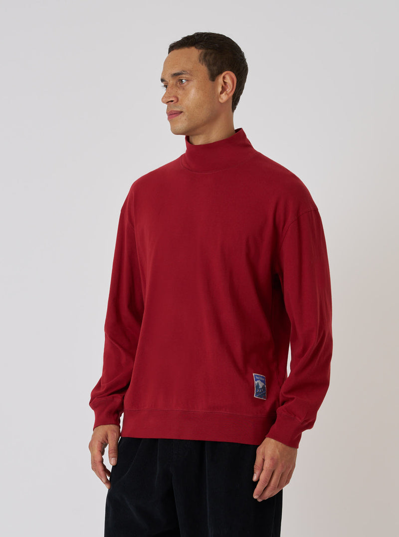 A man poses in a Red Single Jersey Funnel Neck Tee against a white backdrop. The tee has long sleeves and a relaxed fit. He wears dark pants. The image is well-lit.