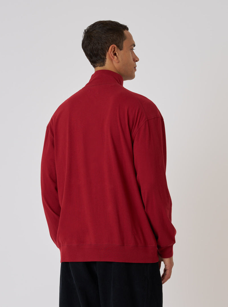 A man stands against a white backdrop, modeling the Red Single Jersey Funnel Neck Tee. The tee has long sleeves and a relaxed fit. He wears dark pants.