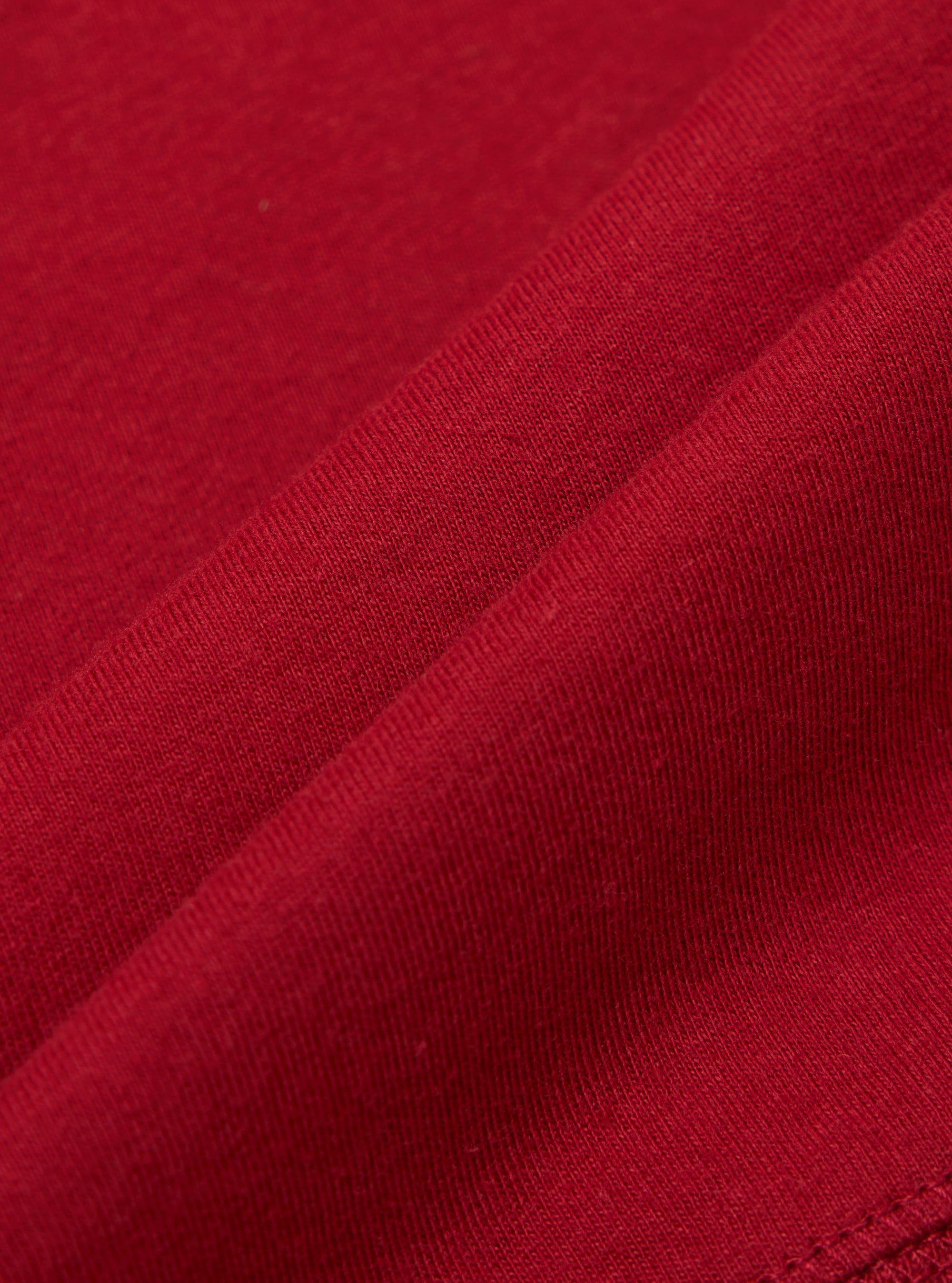 Close-up of the Red Single Jersey Funnel Neck Tee fabric. The soft, red jersey material is draped to show its texture and quality. The image highlights the tee's comfortable fit.
