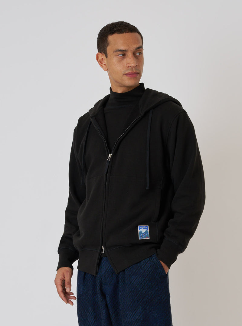 A model poses in front of a white wall wearing a Black Brush Back Sweat New York Hoody. The hoody is black and made of a soft, brushed fabric.