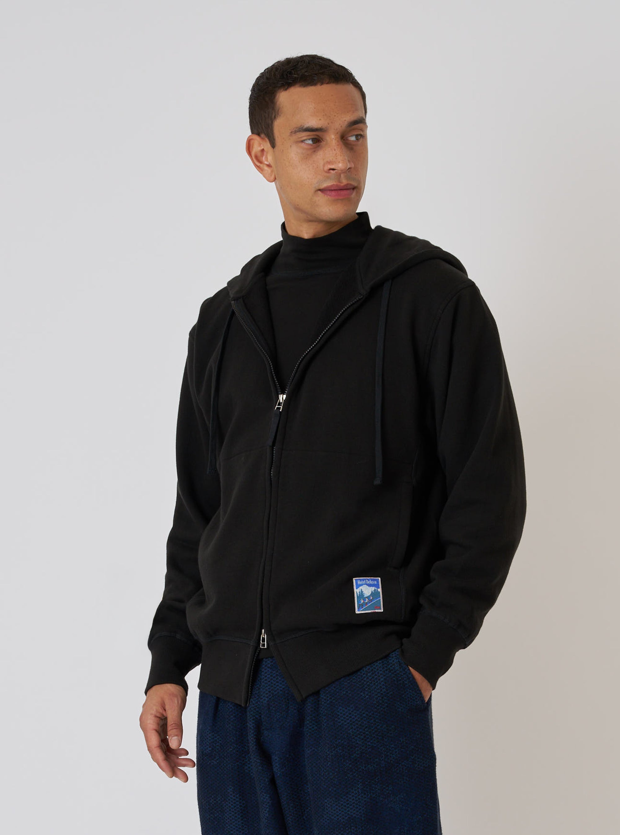 A model poses in front of a white wall wearing a Black Brush Back Sweat New York Hoody. The hoody is black and made of a soft, brushed fabric.