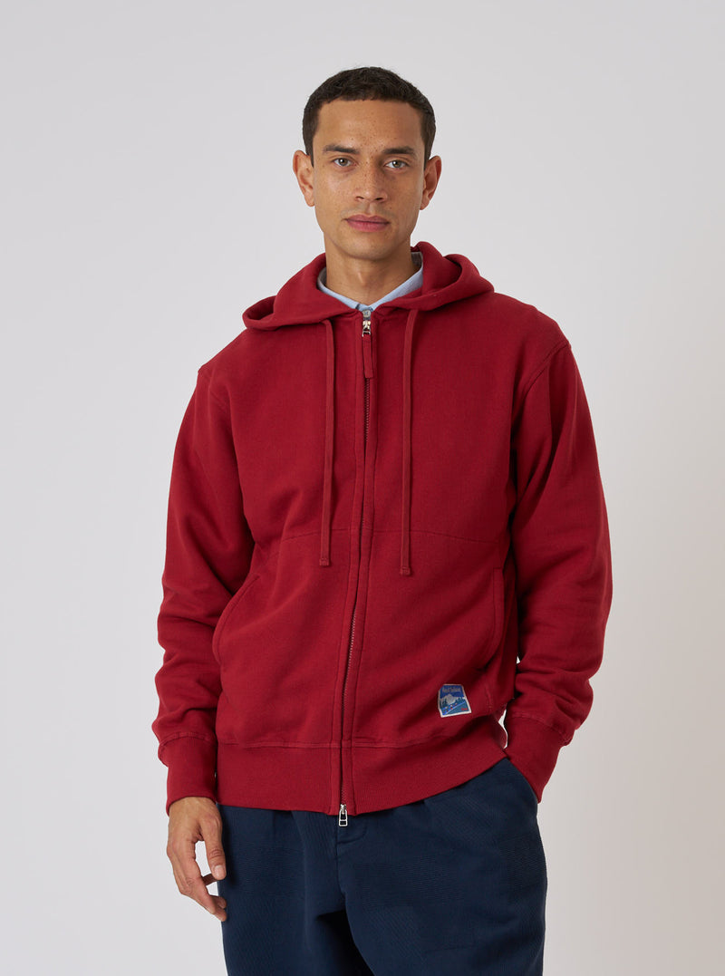 A model poses in front of a white background wearing a Red Brush Back Sweat New York Hoody. The hoody is zipped up and paired with navy trousers.