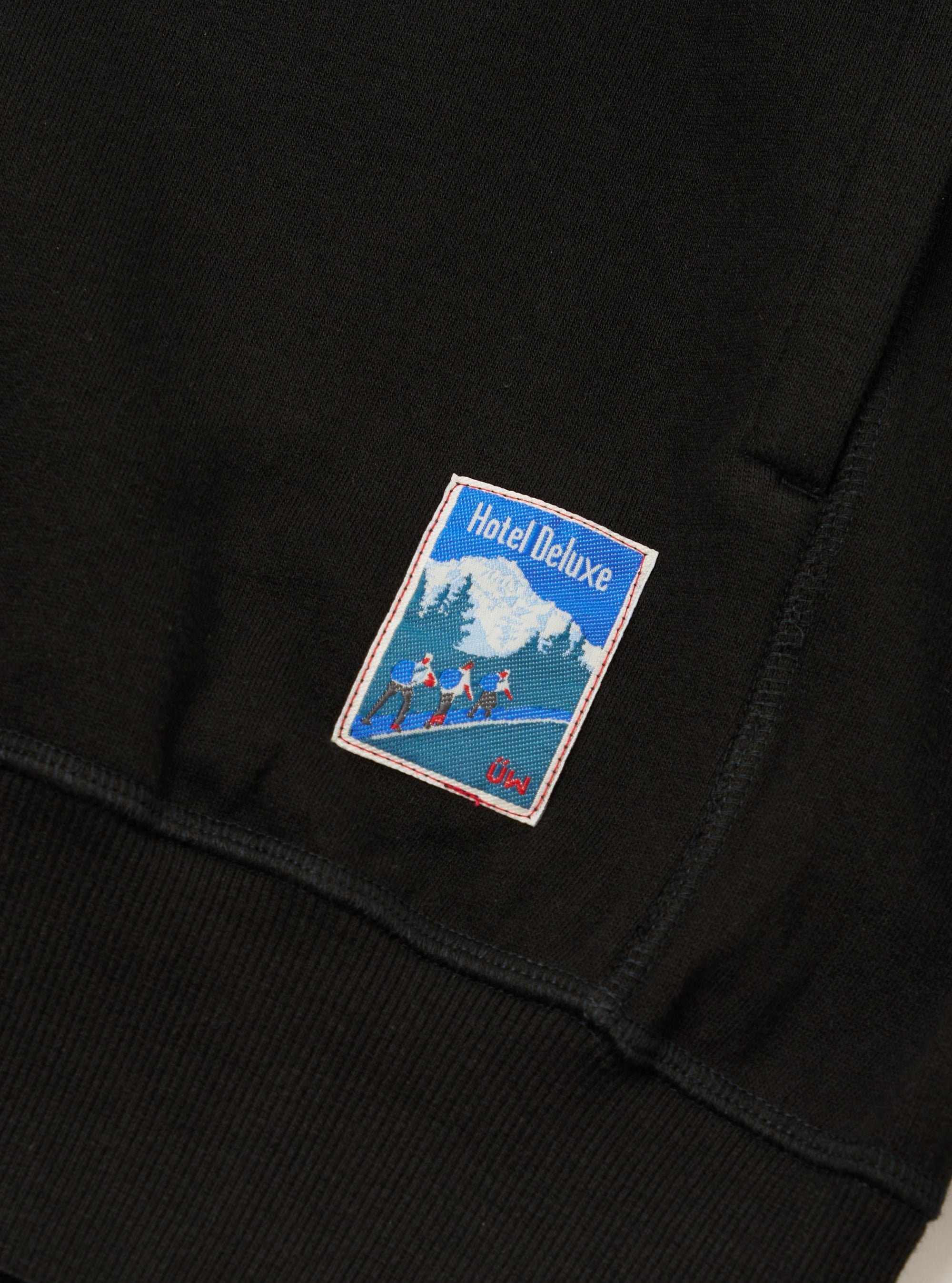 Close-up of the Black Brush Back Sweat New York Hoody, showing the fabric texture and a 'Hotel Deluxe' patch. The image is tightly cropped, focusing on the details.