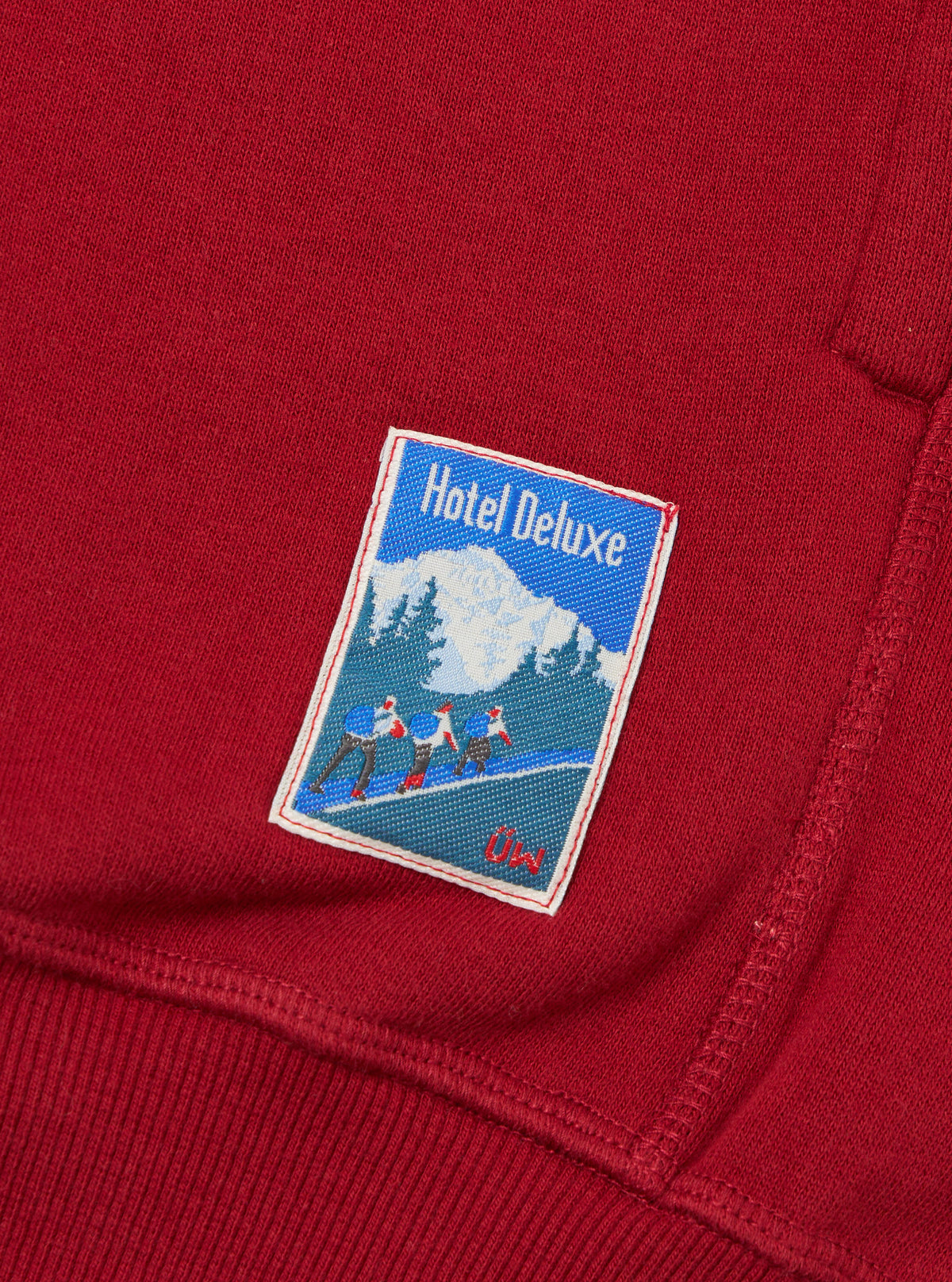 Close-up of the Red Brush Back Sweat New York Hoody showing the fabric texture and a Hotel Deluxe patch with a winter scene. The hoody is laid flat on a neutral surface.