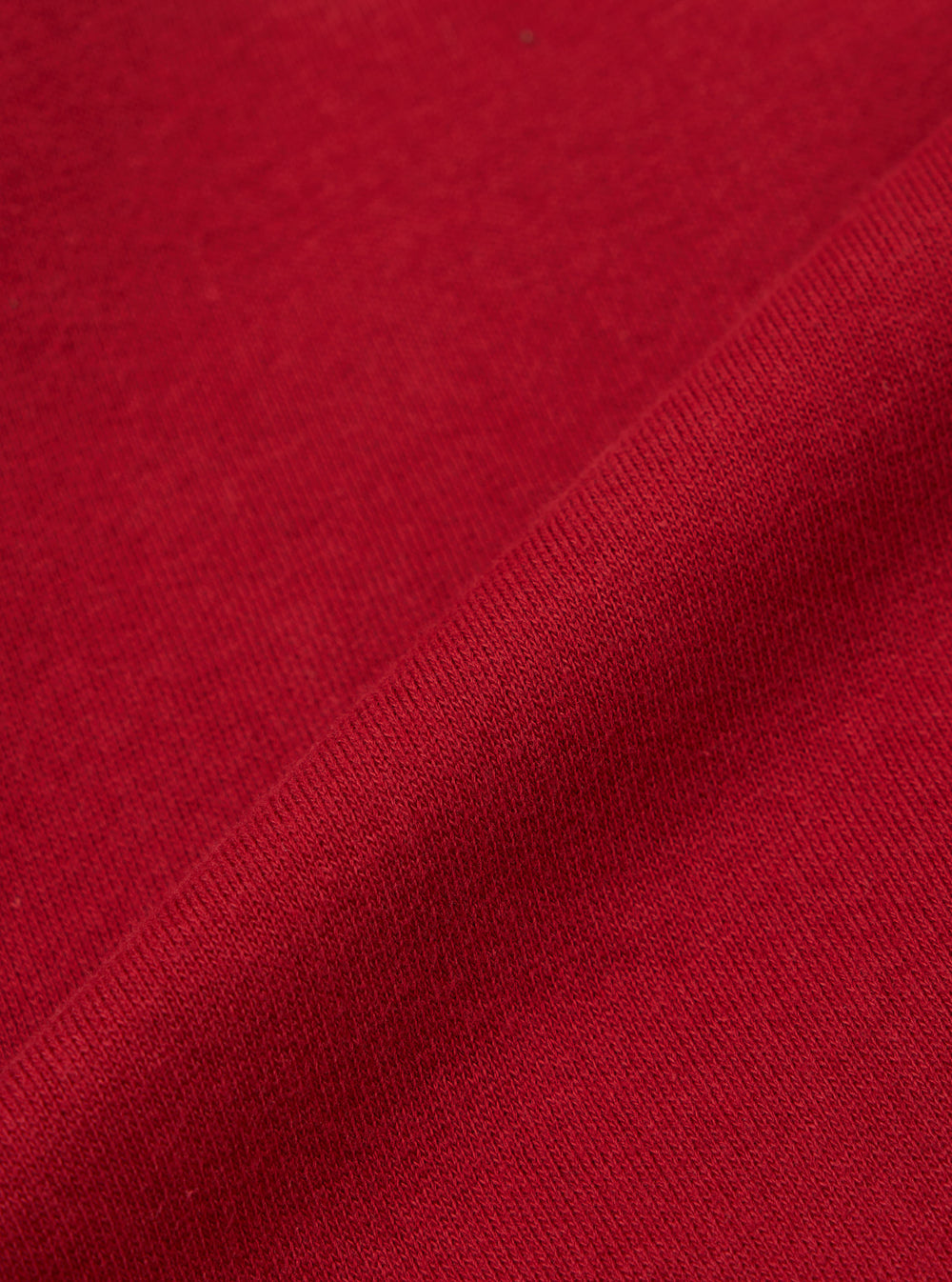 Close-up of the red, textured fabric of the Red Brush Back Sweat New York Hoody. The material is folded, showing the weave. Background is a plain surface.