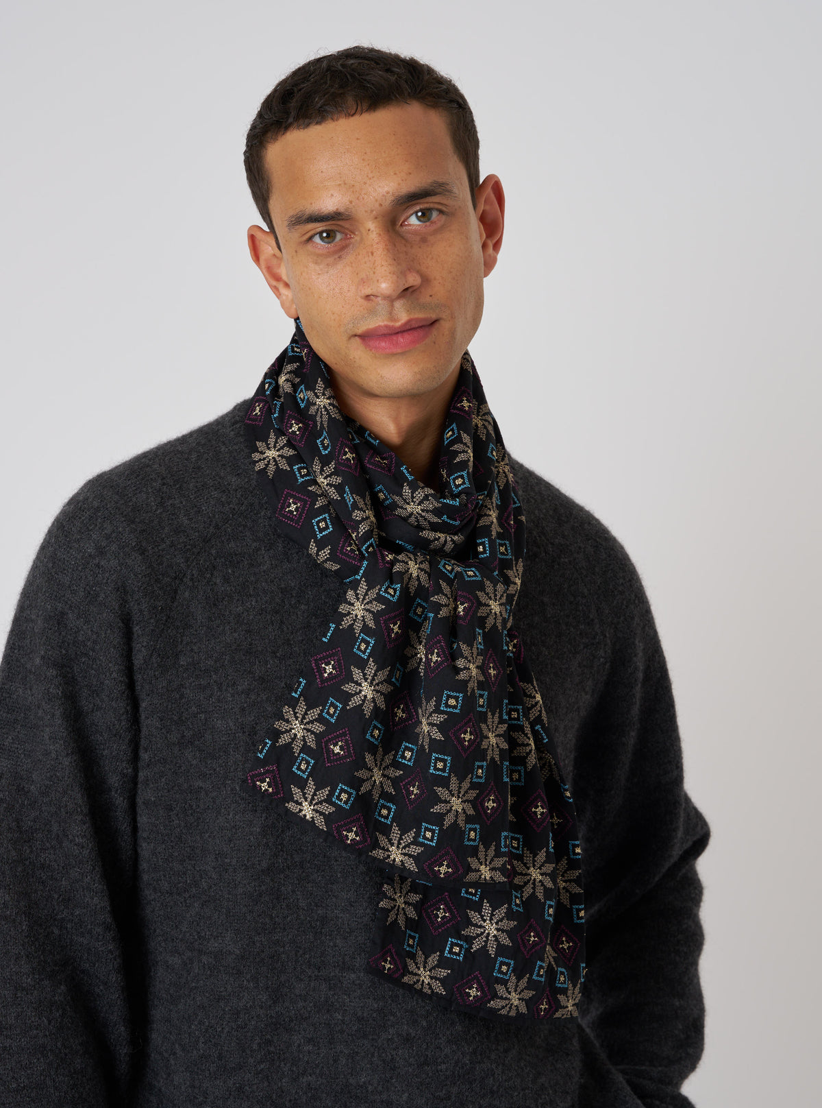 A man in front of a white background wears a Black Embroidery Cotton Satin Embroidery Scarf. The scarf has gold, blue, and purple embroidery.