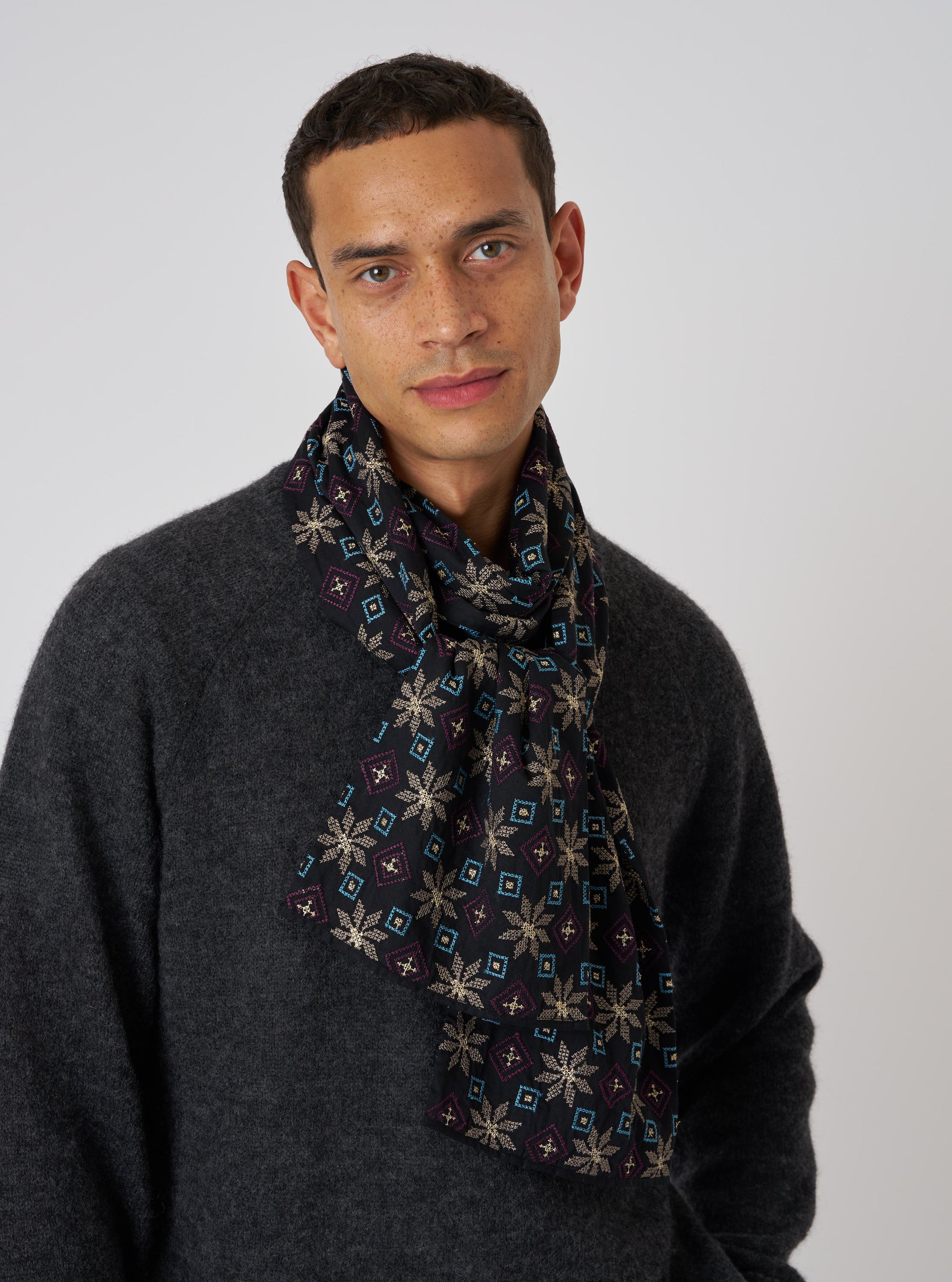 A man in front of a white background wears a Black Embroidery Cotton Satin Embroidery Scarf. The scarf has gold, blue, and purple embroidery.
