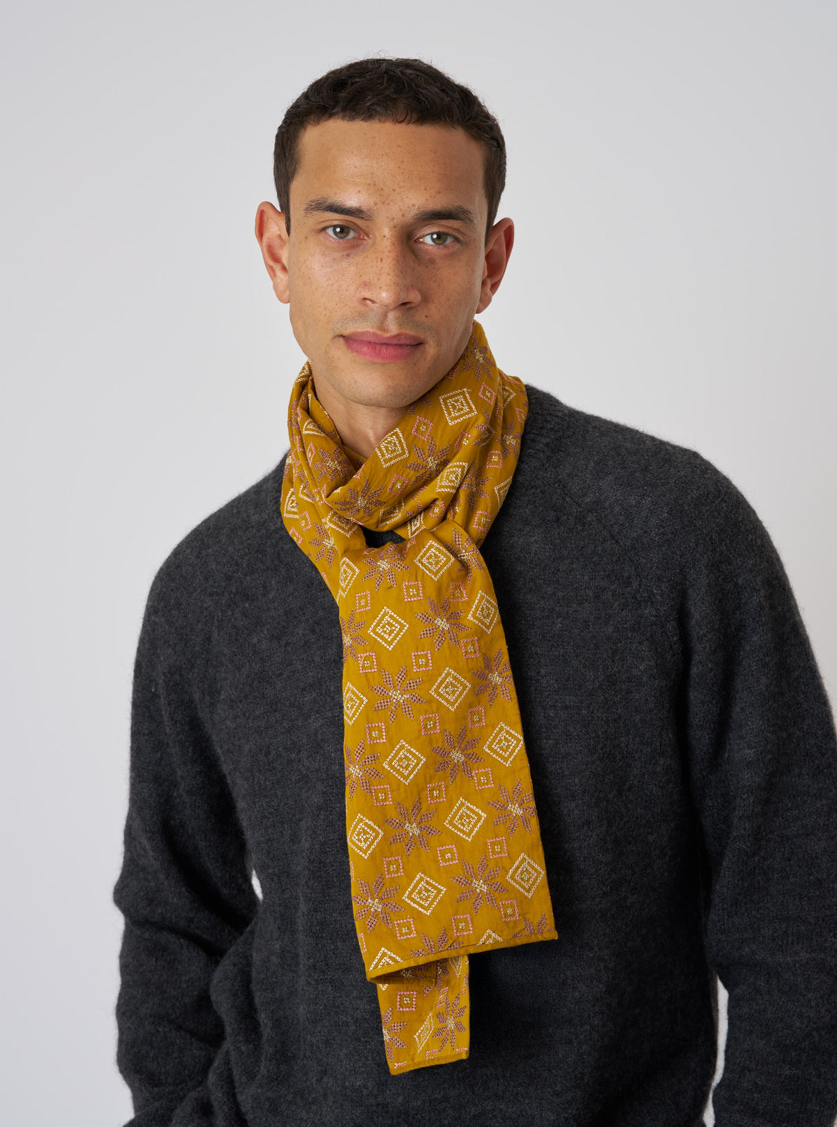 A man models the Mustard Embroidery Cotton Satin Embroidery Scarf. The mustard scarf has a geometric pattern. He wears a gray sweater against a white backdrop.