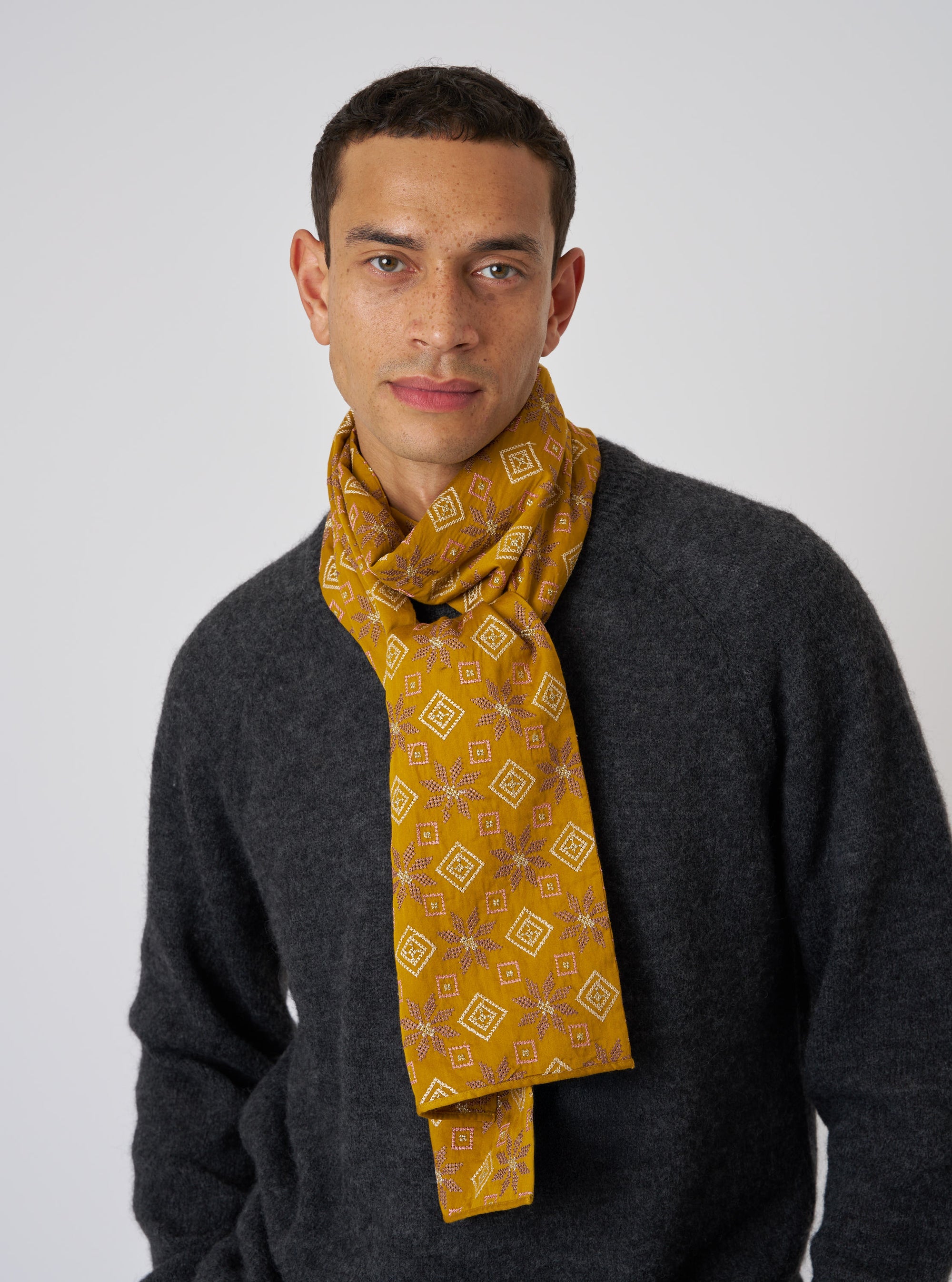 A man models the Mustard Embroidery Cotton Satin Embroidery Scarf. The mustard scarf has a geometric pattern. He wears a gray sweater against a white backdrop.