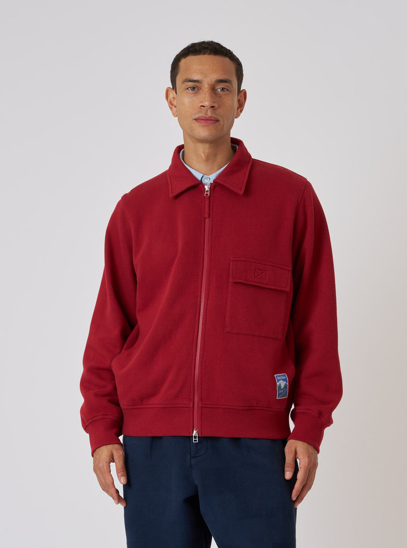 A model poses in a Red Brush Back Sweat Tyler Sweat Jacket against a white background. The jacket is paired with navy pants. The jacket features a zip front and chest pocket.