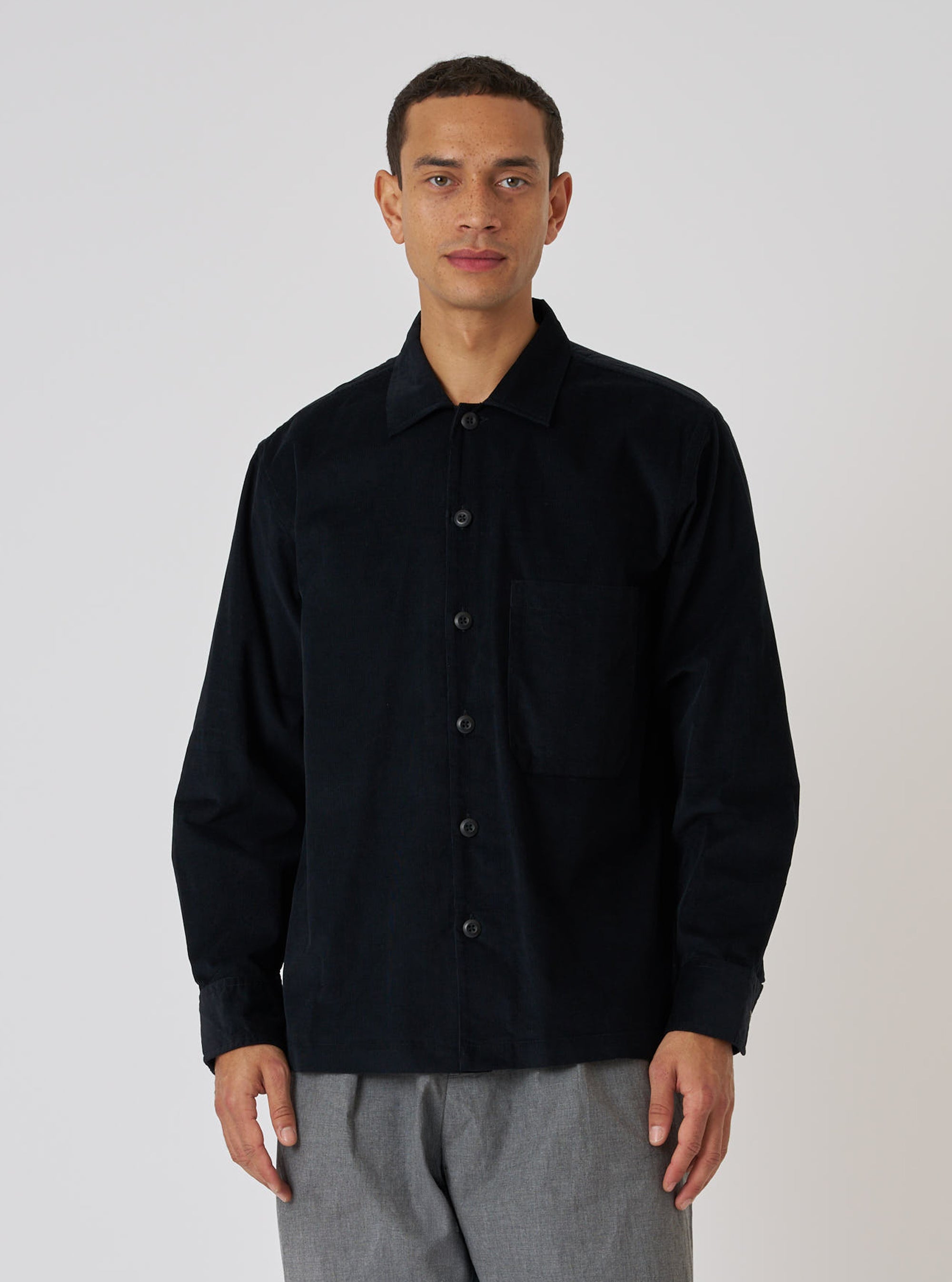A model wears a Black Organic Fine Cord Fatigue Shirt against a white backdrop. The shirt is buttoned and paired with gray pants.