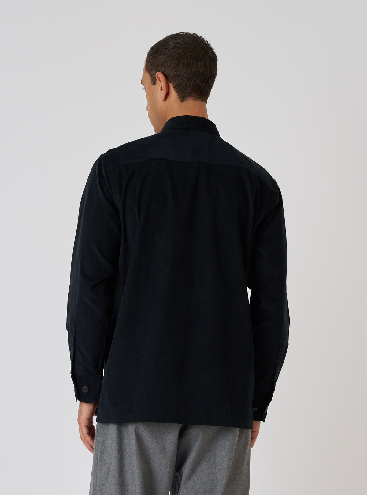 Back view of a model wearing a Black Organic Fine Cord Fatigue Shirt. The shirt is paired with gray pants, set against a clean white backdrop.
