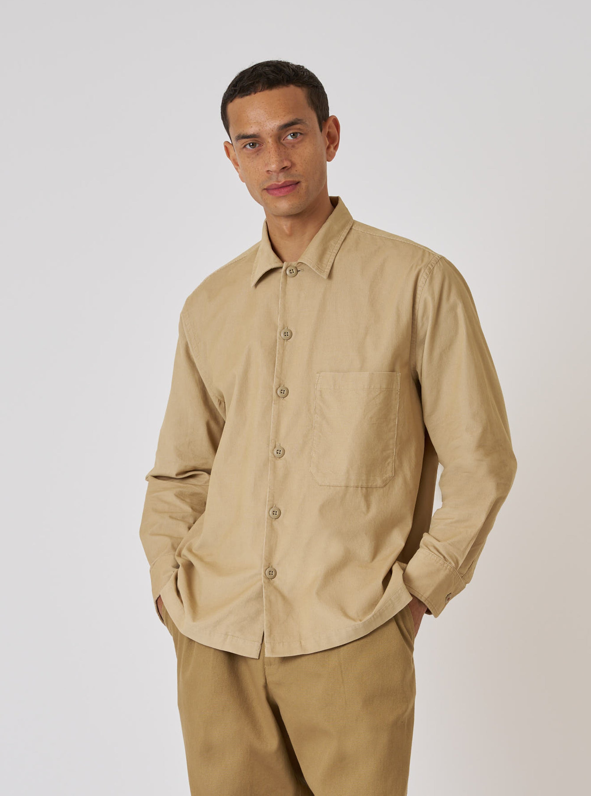 A model poses in a Sand Organic Fine Cord Fatigue Shirt. The shirt is a light tan color and made of corduroy. He is wearing matching pants, standing against a plain white background.