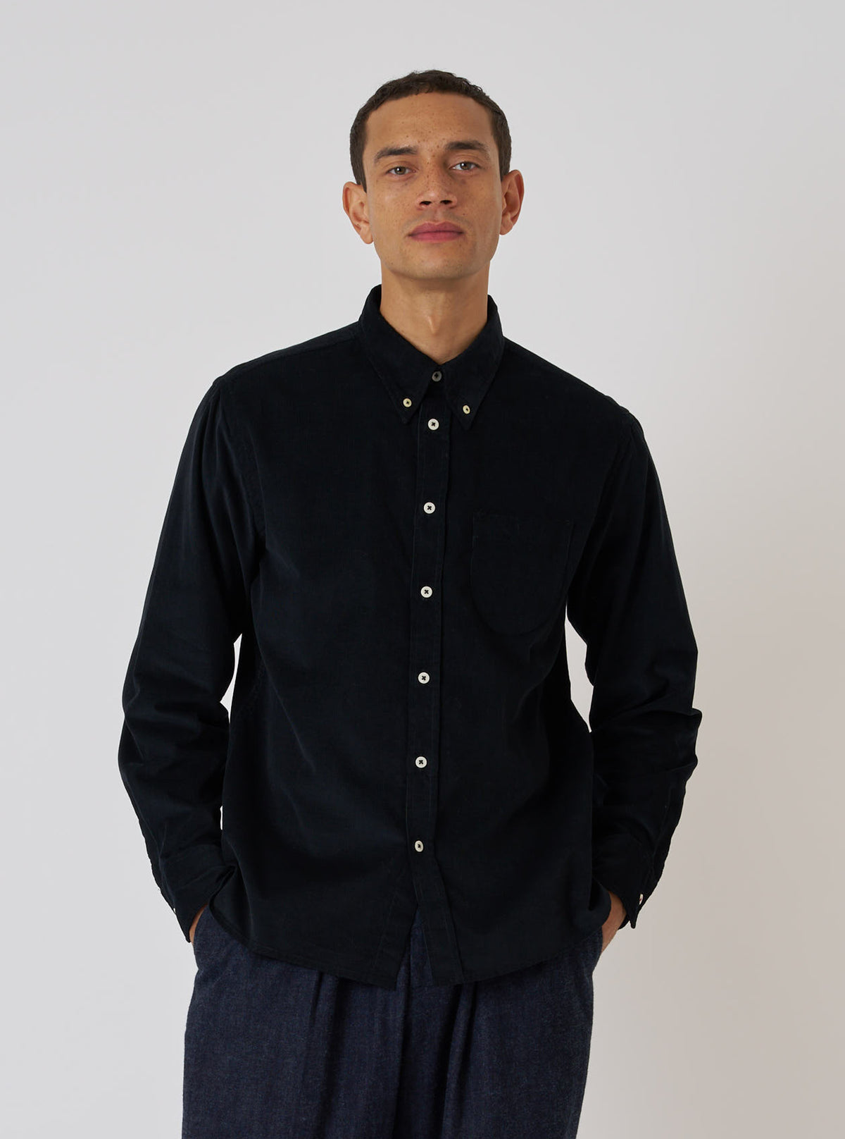 A model poses in front of a white background, wearing a Black Organic Fine Cord Daybrook Shirt. The shirt is buttoned and has long sleeves.