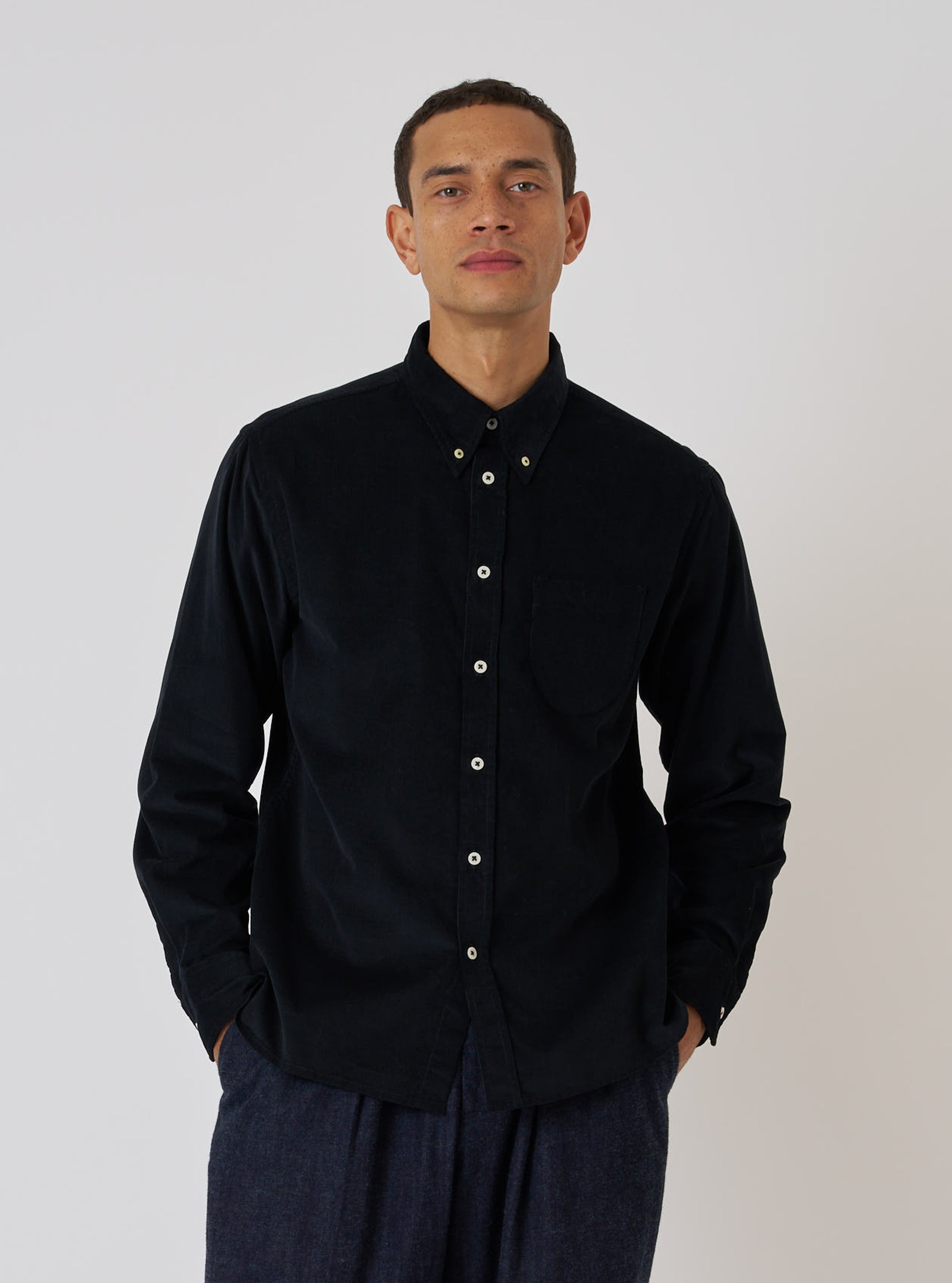 A model poses in front of a white background, wearing a Black Organic Fine Cord Daybrook Shirt. The shirt is buttoned and has long sleeves.