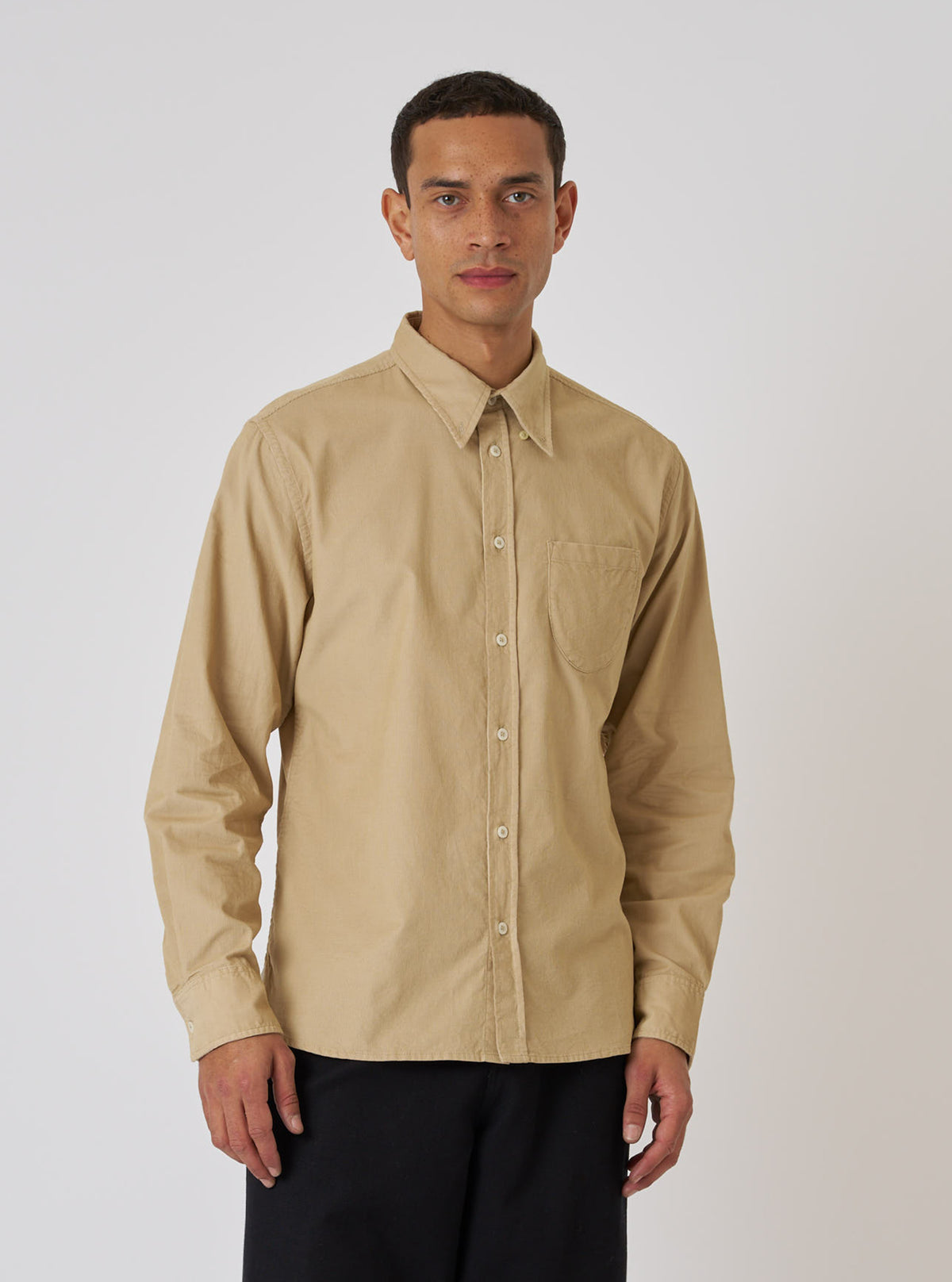 A model poses in front of a white background wearing the Sand Organic Fine Cord Daybrook Shirt. The shirt is a light tan color and has a relaxed fit.