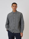 A man models the Charcoal Organic Chambray Patched Lazy Day Shirt. The gray shirt has a button-down collar and chest pocket. He wears dark pants. White backdrop.