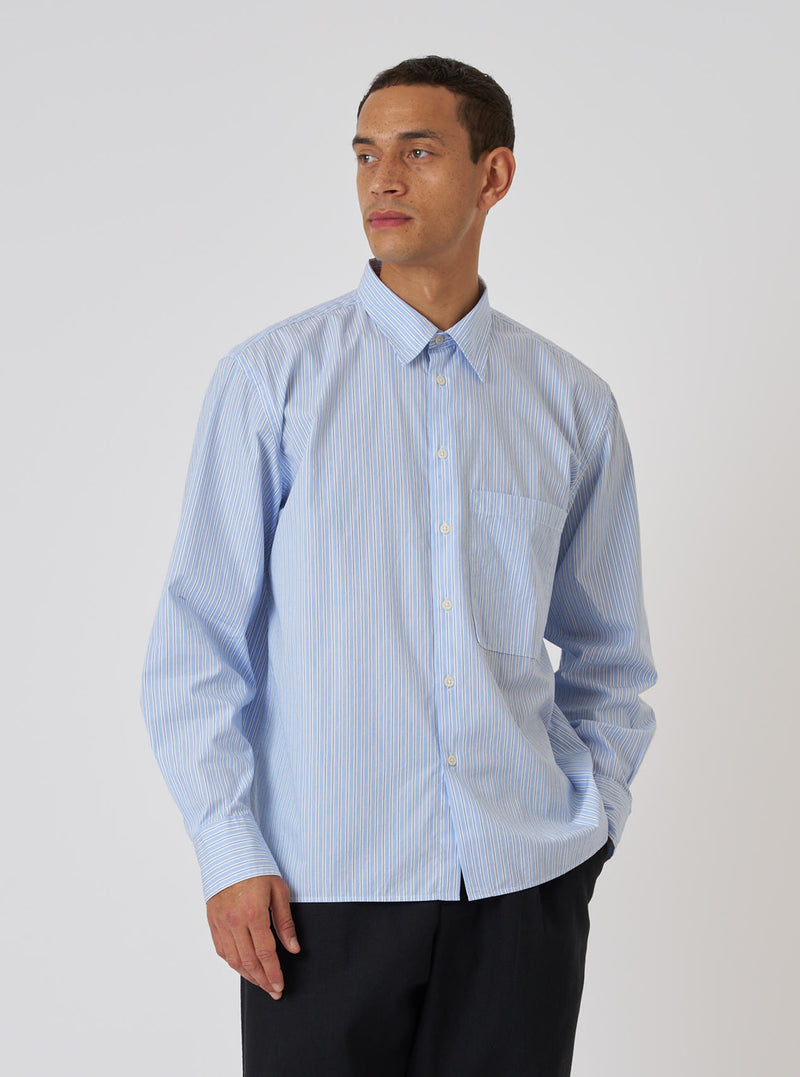 A man poses in a Blue Office Stripe Square Pocket Shirt against a white backdrop. The shirt is blue with white stripes and has a square pocket. He wears black pants.