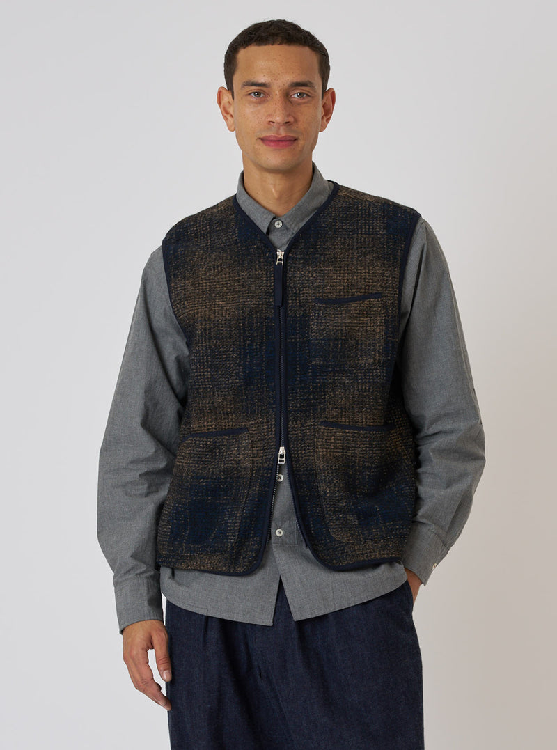 A model poses in front of a plain backdrop, wearing a Navy Deca Check Zip Gilet. The gilet features a navy and brown check pattern and a zip-up front.