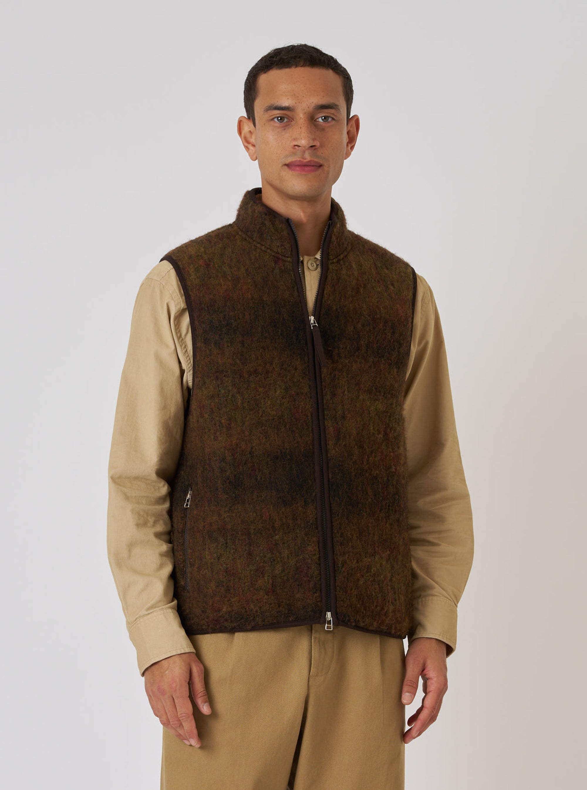 A model poses in a Brown Alps Fleece Climbing Gilet. The fleece is brown with a full zip. He wears a tan shirt and trousers. Shot in a studio on a white backdrop.