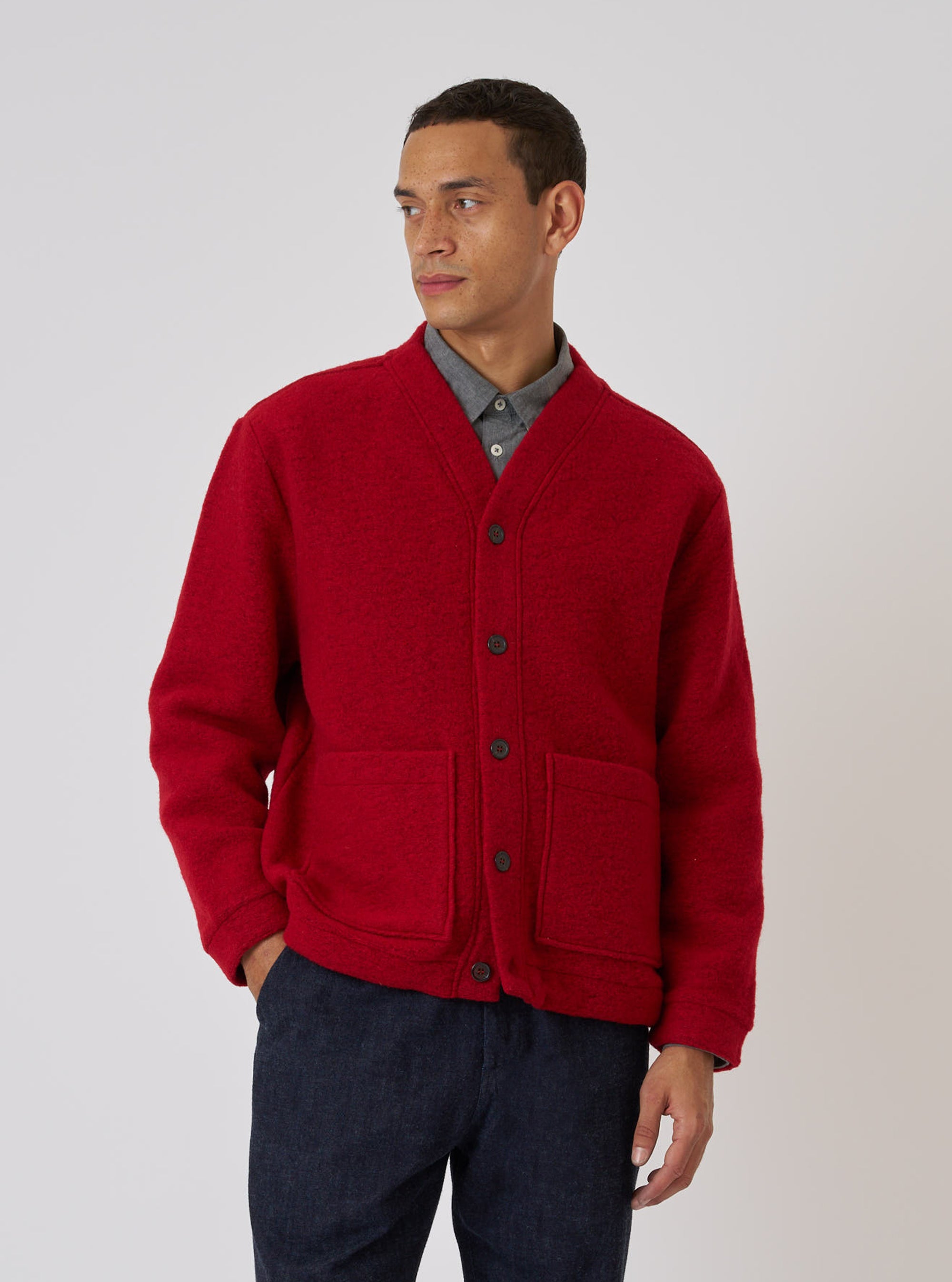 A man models the Red Wool Fleece Advance Cardigan against a plain background. The red, button-up cardigan has patch pockets. He wears a gray shirt and dark pants.
