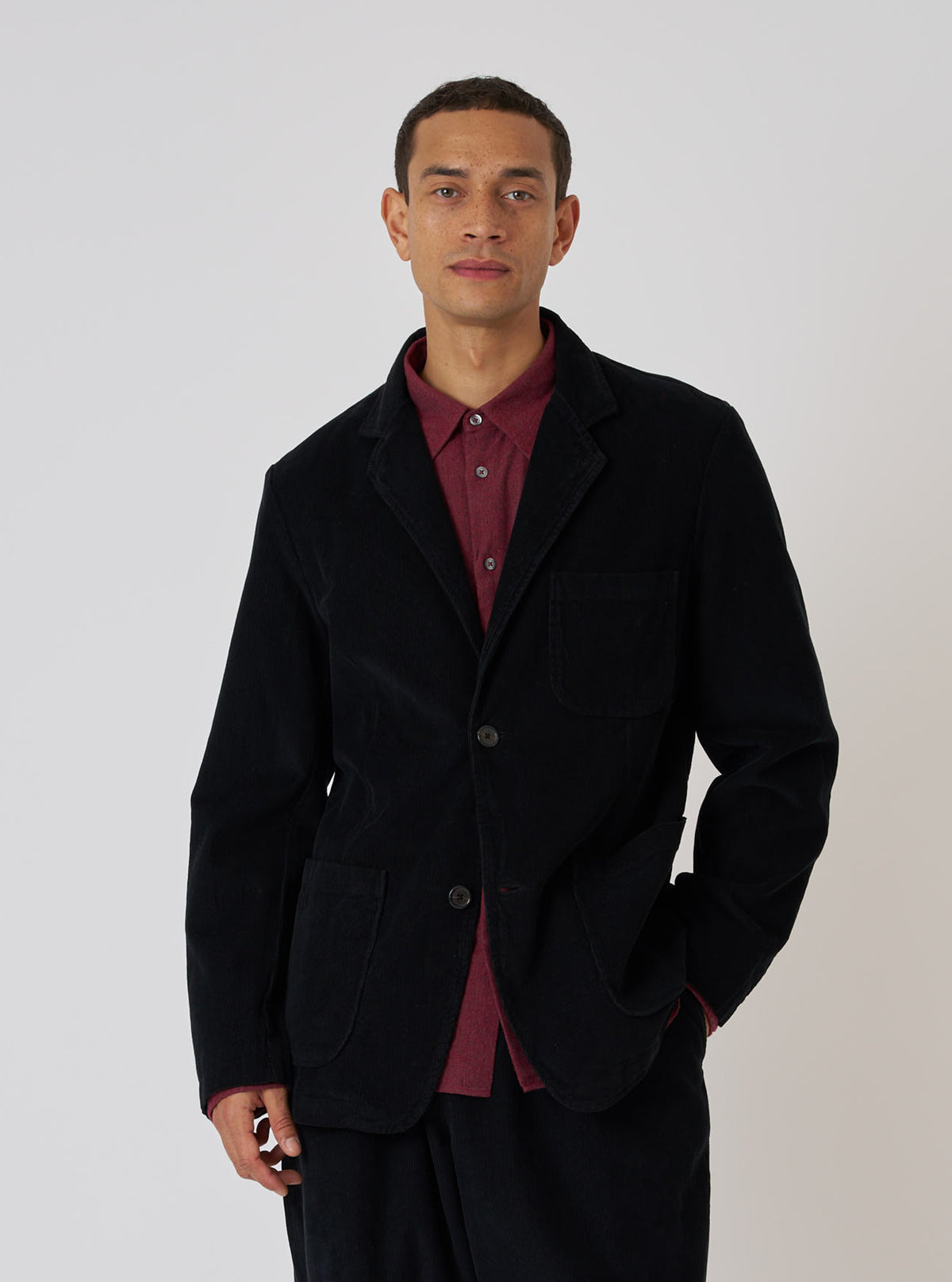 A model poses in a Black Cat Cord Two Button Jacket. The jacket is black corduroy, worn over a red shirt. The background is a plain white studio backdrop.