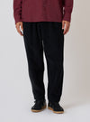 A person stands against a white backdrop, showcasing the Black Cat Cord Pleated Track Pant. The black cord pants are paired with a maroon shirt and black sneakers.