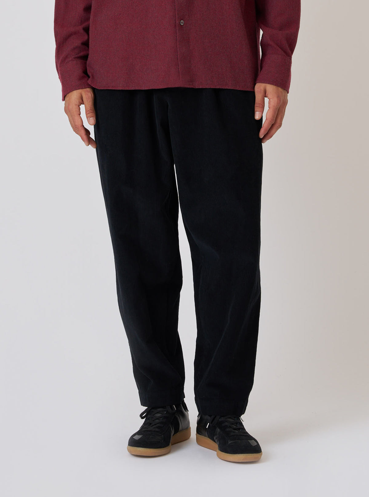 A person stands against a white backdrop, showcasing the Black Cat Cord Pleated Track Pant. The black cord pants are paired with a maroon shirt and black sneakers.