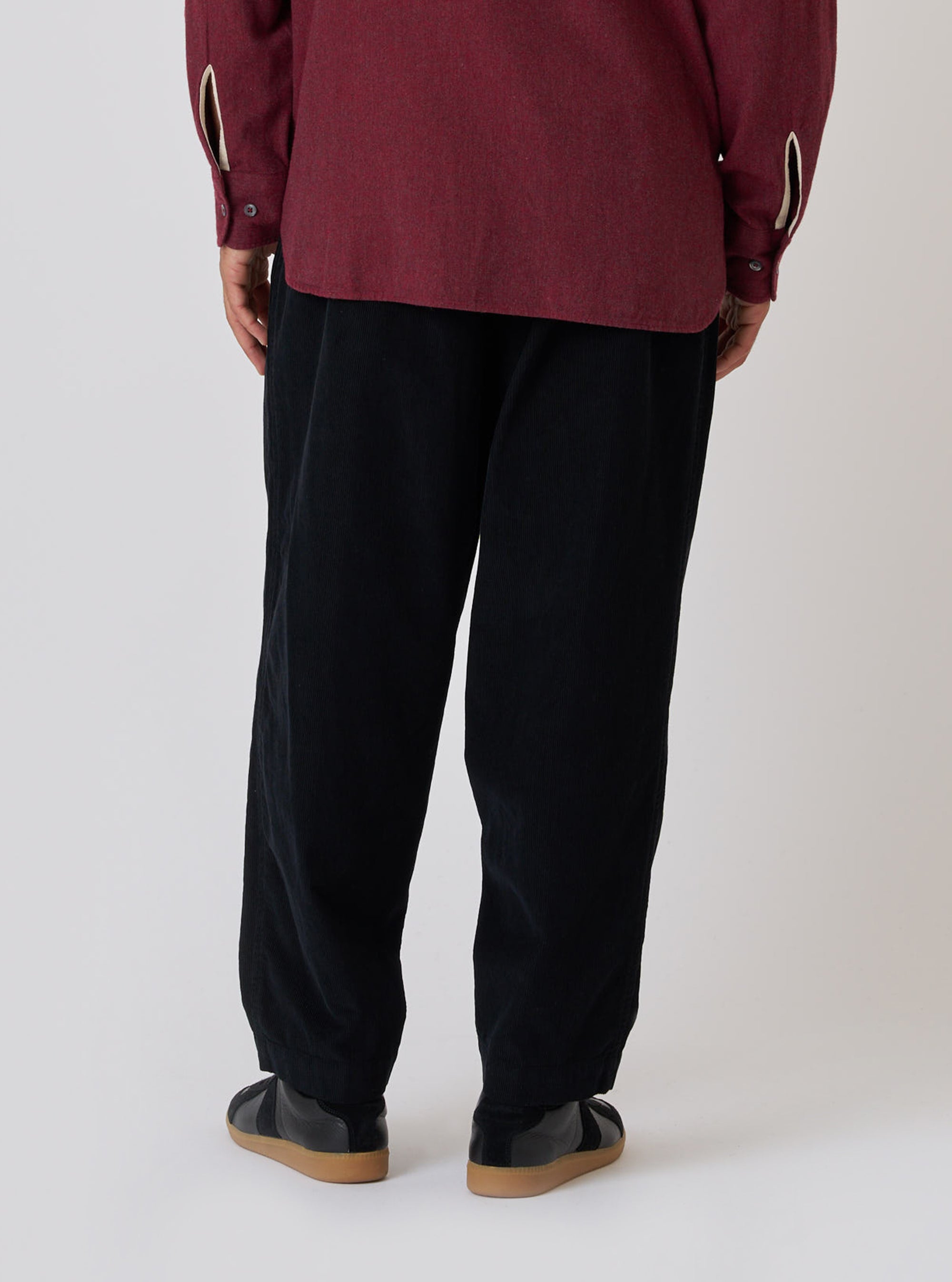 Back view of a model wearing the Black Cat Cord Pleated Track Pant. The black corduroy pants are paired with a maroon shirt and black shoes. Simple studio background.