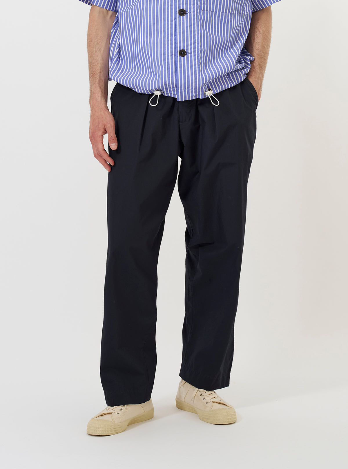 A model is wearing the Navy Broadcloth Oxford Pant against a plain white background. The navy blue trousers are shown from the front, featuring a loose, wide-leg fit, a pleated front, and a drawstring waistband. The model is standing and wearing the pants with a casual shirt and shoes.