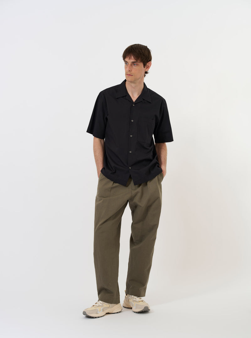 The Black Broadcloth Camp Shirt is shown laid flat against a solid white background. The shirt features a camp collar, a full button-down front, and short sleeves. A single patch pocket is visible on the left chest. The garment is solid black with a smooth, matte finish.