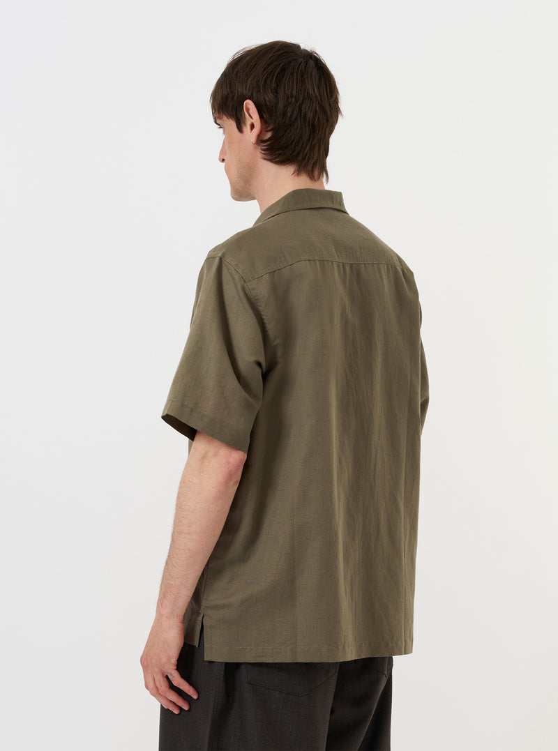 A model is wearing the Olive Linen Cotton Shirting Camp Shirt against a plain white background. The image shows the shirt from the back, highlighting the yoke with small pleats and the elongated hem with side splits. The model is standing with their hands in their pockets, and the olive green shirt is paired with light-colored trousers.