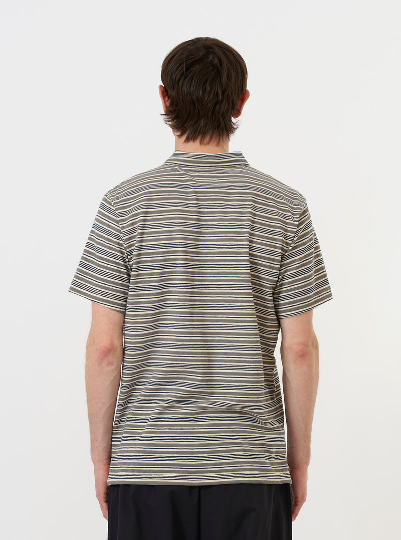 A model stands against a plain white background wearing the Grey OS Stripe Jersey Vacation Polo. The image shows the garment from the back, highlighting the grey and white horizontal stripes, the straight hem, and the relaxed fit of the short-sleeved polo shirt.