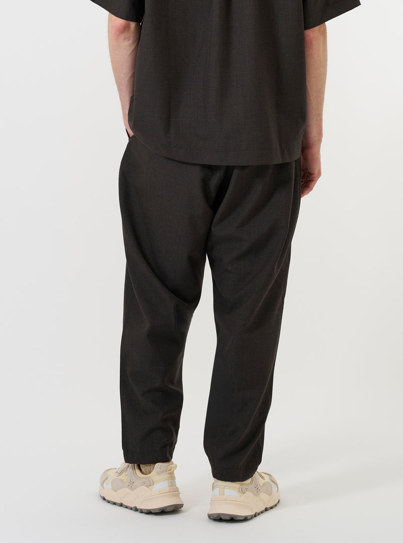 A model is wearing the Brown Marl Tropical Suiting Pleated Track Pant against a plain white background. The image, captured from a side-rear perspective, showcases the loose, tapered fit and the brown marl texture of the lightweight tropical suiting fabric. The model is standing, highlighting the drape of the trousers and the narrow hem at the ankle.