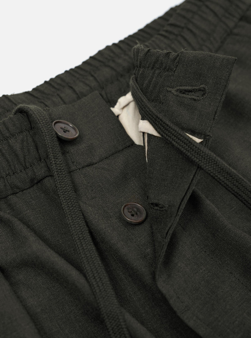 A pair of Olive Marl Tropical Suiting Pleated Track Pants is shown from the back, laid flat against a plain white background. The trousers feature a loose, tapered leg and an elasticated waistband. Key details include two distinct rear pockets, one with a buttoned flap and one jetted with a button closure.