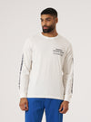 A model wears the Paraboot x Universal Works Ecru Single Jersey Long Sleeve Tee against a white backdrop. The tee is off-white with text on the chest and sleeves.