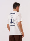 The Paraboot x Universal Works Ecru Single Jersey Print Tee is worn by a model against a plain white background. The image, captured from the rear three-quarter angle, highlights the back of the ecru-colored t-shirt featuring a large dark graphic print of a shoe. The model is also wearing dark trousers and white socks.