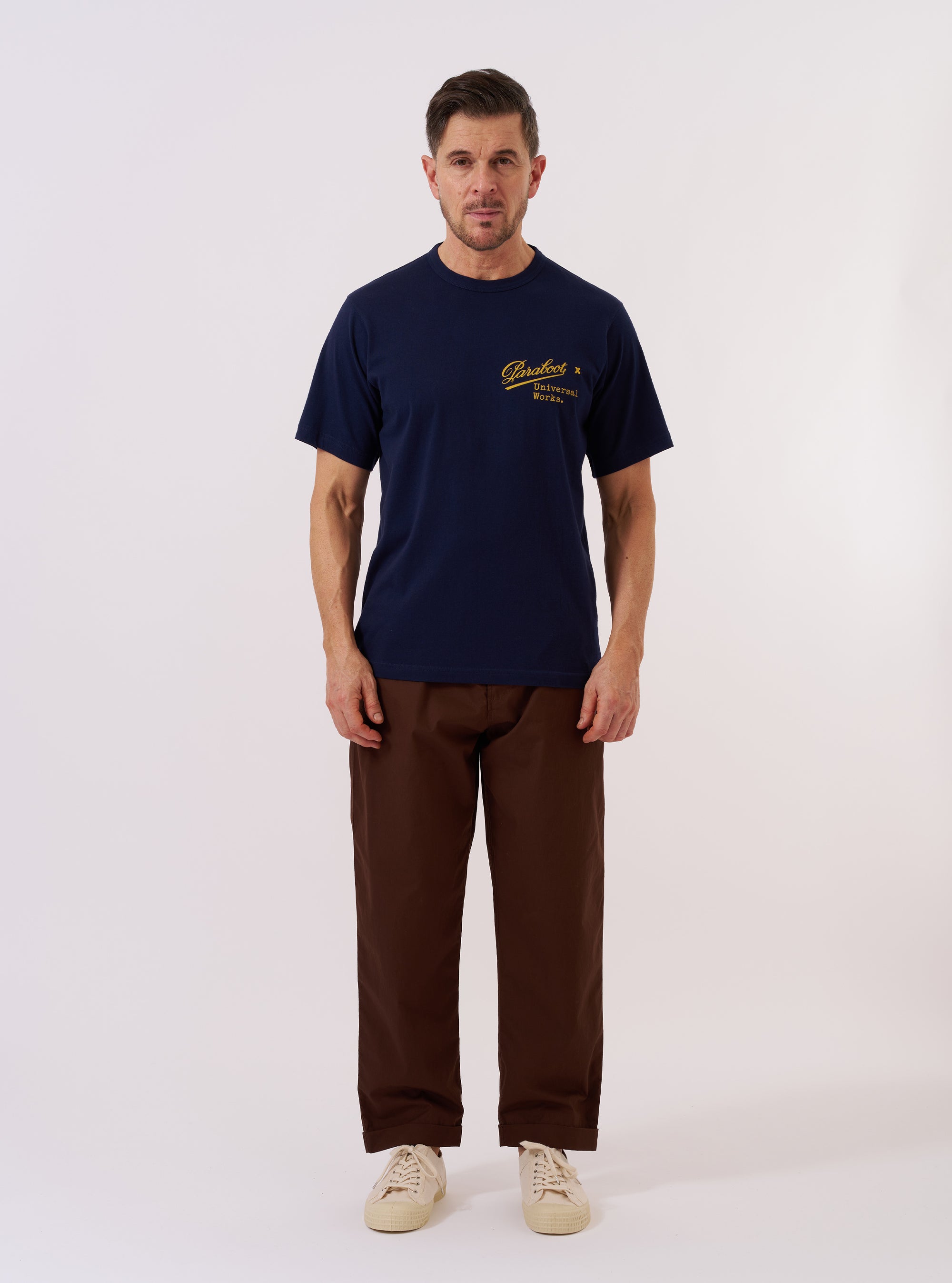 The Paraboot x Universal Works Navy Single Jersey Print Tee is shown as a flat lay against a white background. The short-sleeve t-shirt features a ribbed crewneck and a prominent white graphic print on the front inspired by vintage advertising. The garment is made from a solid navy blue cotton jersey.