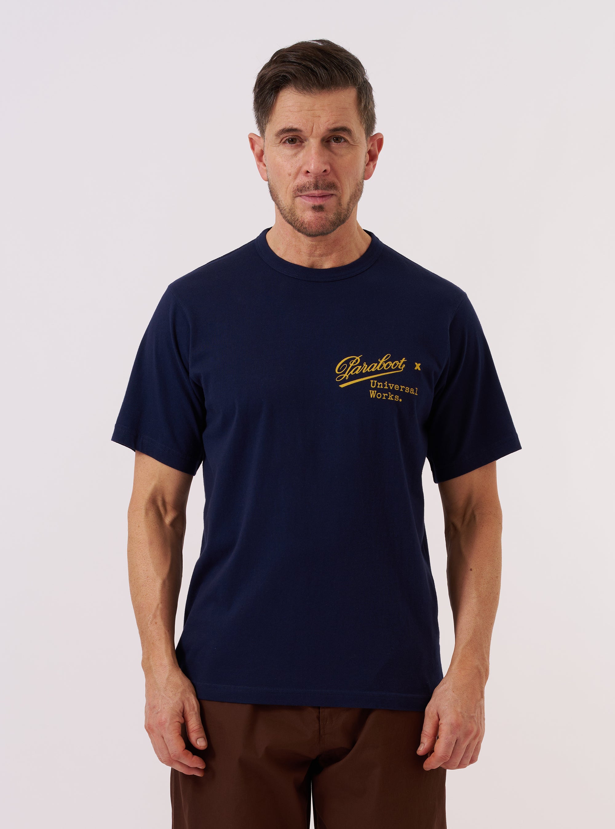 A model wears the Paraboot x Universal Works Navy Single Jersey Print Tee against a plain white background. The navy blue cotton T-shirt features a relaxed fit, a ribbed crew neckline, and a large graphic print on the front inspired by vintage advertising. The model is shown from the front, highlighting the short sleeves and the central placement of the white and light blue graphic.