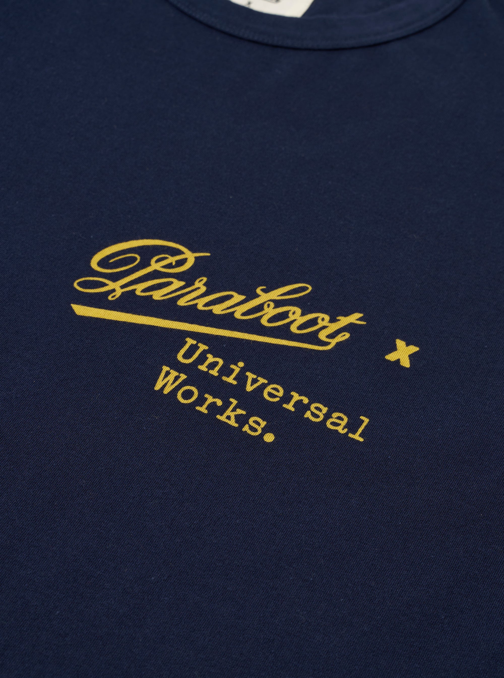 The Paraboot x Universal Works Navy Single Jersey Print Tee is shown from a rear three-quarter angle as a flat lay against a plain white background. The image highlights the relaxed fit of the navy blue cotton t-shirt and features a large white graphic print on the back. The ribbed crew neck and short sleeves are clearly visible.