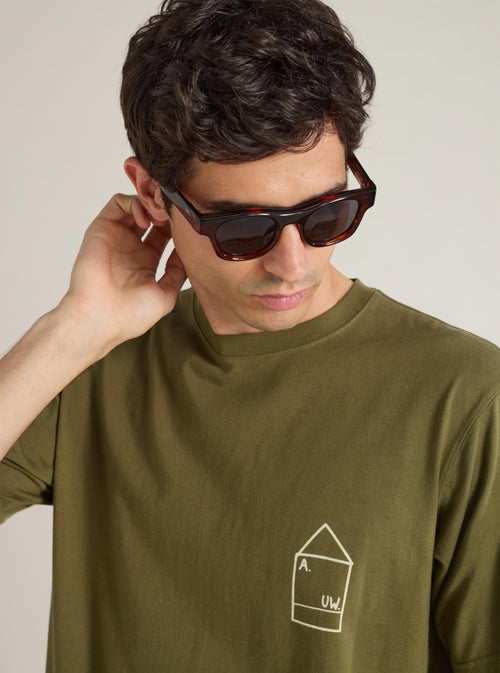 A model wears A. SOCIETY x Universal Works Brown Stanley Sunglasses. The brown frames complement his olive tee. Shot against a neutral backdrop.