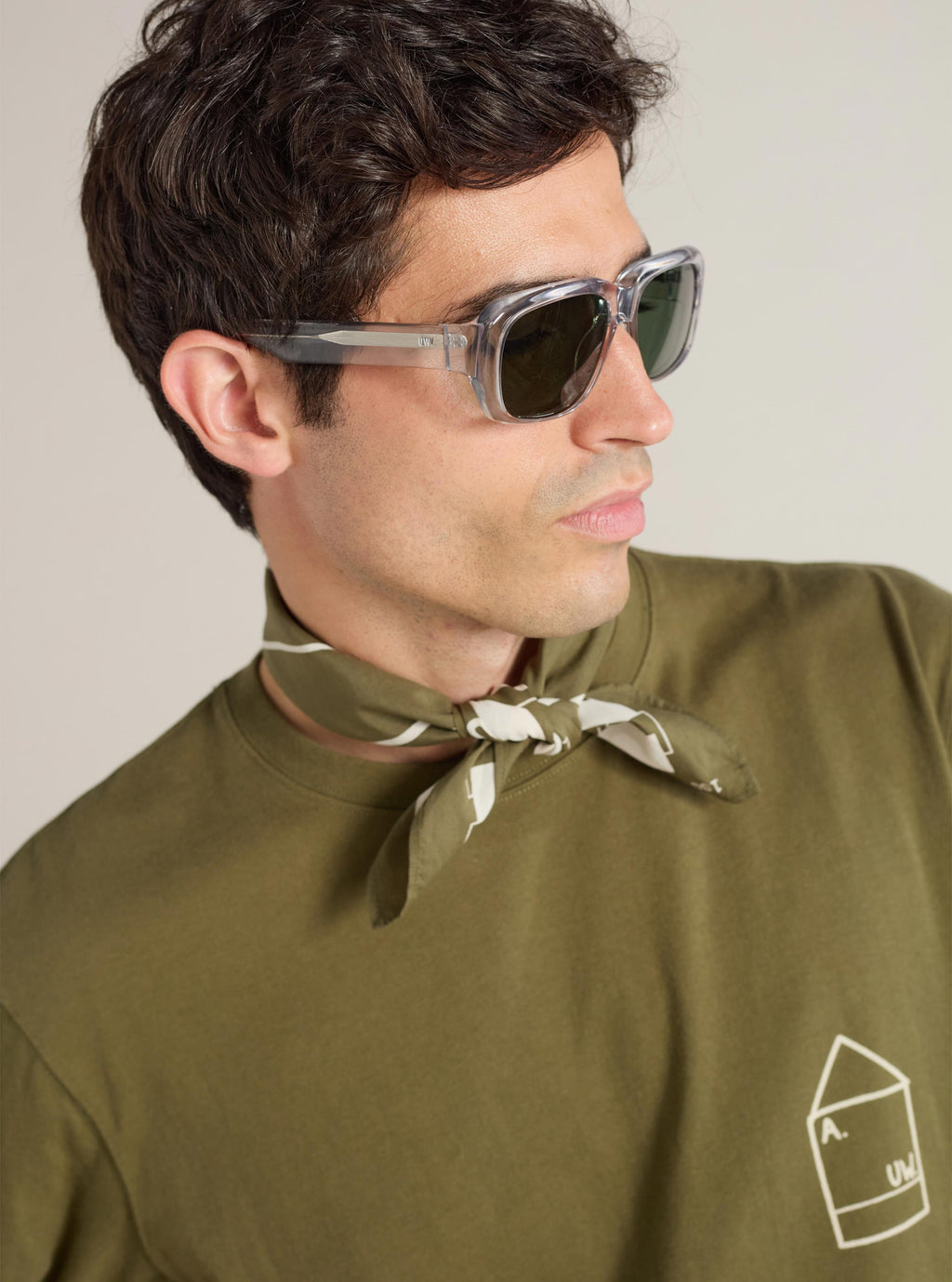 Profile of a model wearing A. SOCIETY x Universal Works Grey Aberdeen Sunglasses. The grey frame complements his olive tee and neck scarf, set against a neutral background.
