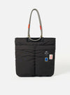 The Topologie x Universal Works Black UW Tech Helmet Bag, a black padded tote with rope handles, stands on a white background. Patches adorn the front.