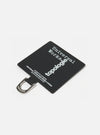 Close-up of the Topologie x Universal Works Black UW Tech Phone Strap Adapter on a white surface. The black fabric adapter features white text and a silver metal loop.
