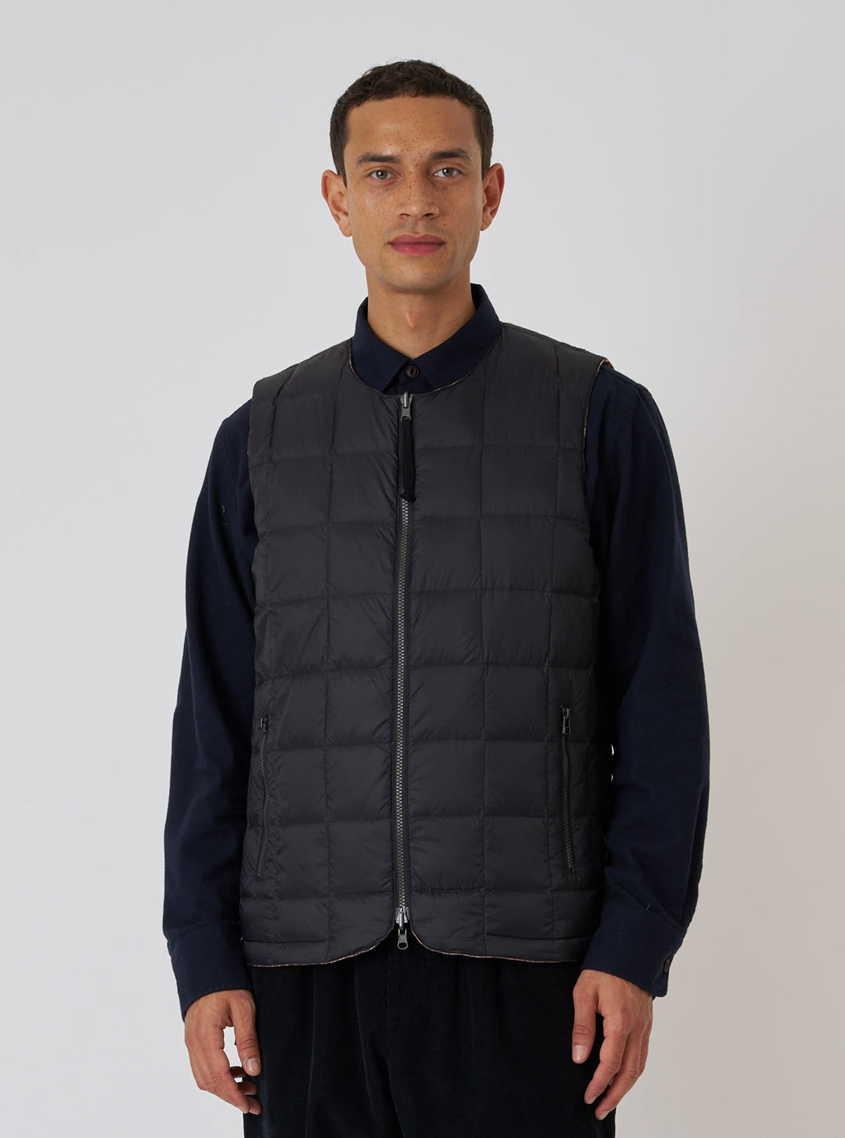 A man stands against a white backdrop, modeling the TAION x Universal Works Reversible Black Military Line Vest. The vest is black, quilted, and zipped up, worn over a navy shirt.