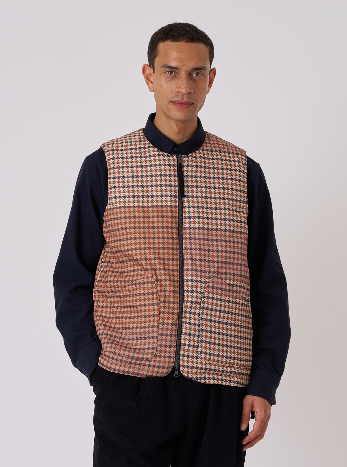 A model poses in the TAION x Universal Works Reversible Black Military Line Vest. The vest is brown plaid with a black zipper. He wears a navy shirt and black pants against a white backdrop.