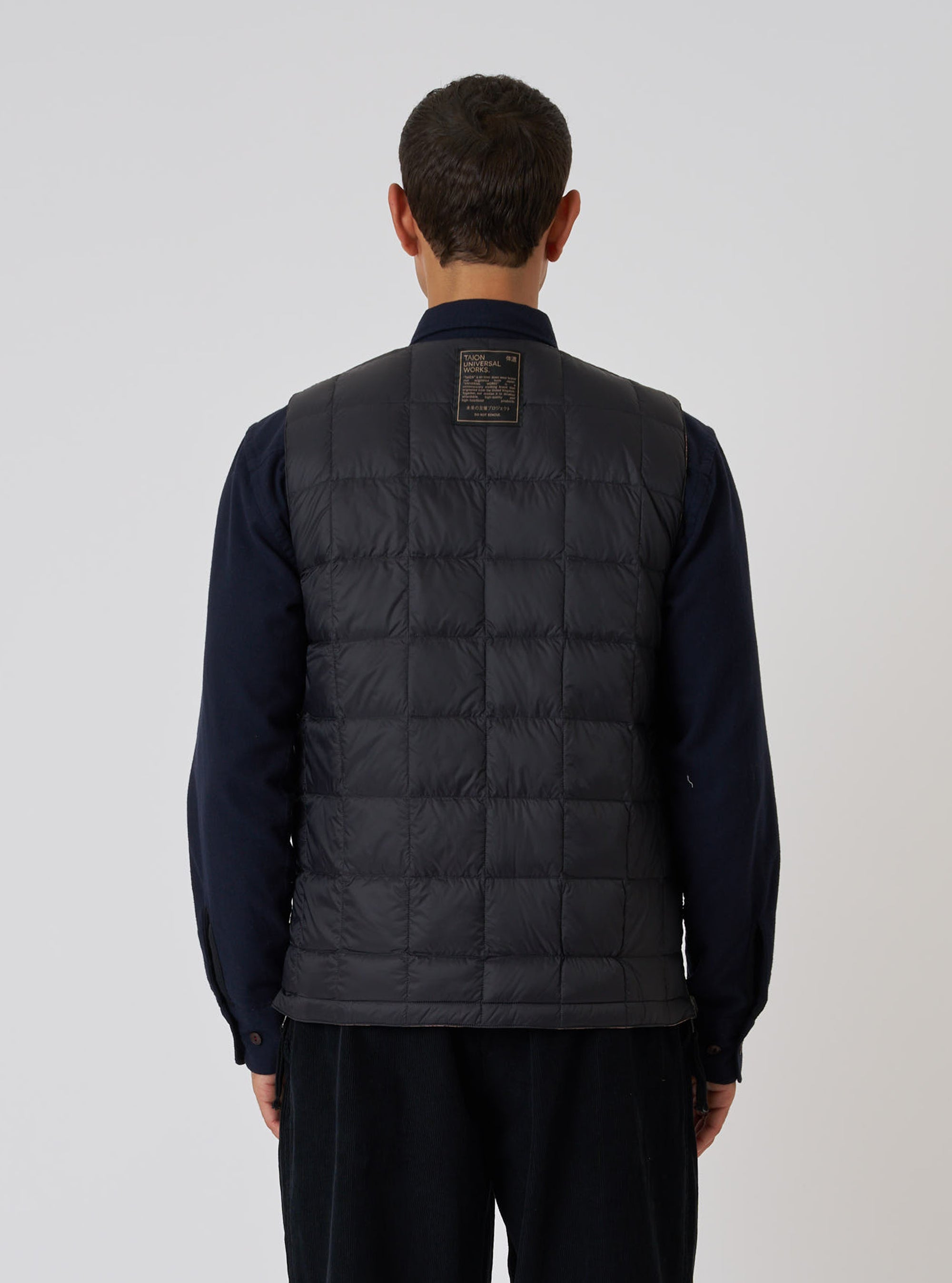 Back view of a model wearing the TAION x Universal Works Reversible Black Military Line Vest. The vest is black with a quilted pattern. Shot against a plain white background.