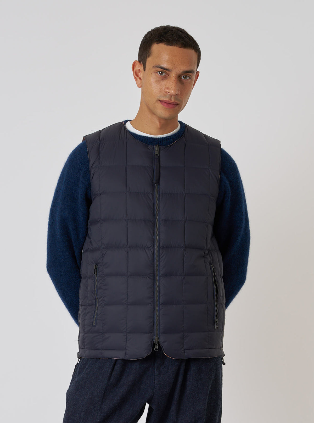 A man models the TAION x Universal Works Reversible Dark Navy Military Line Vest against a white backdrop. The vest is quilted and dark navy. He wears a blue sweater and dark pants.